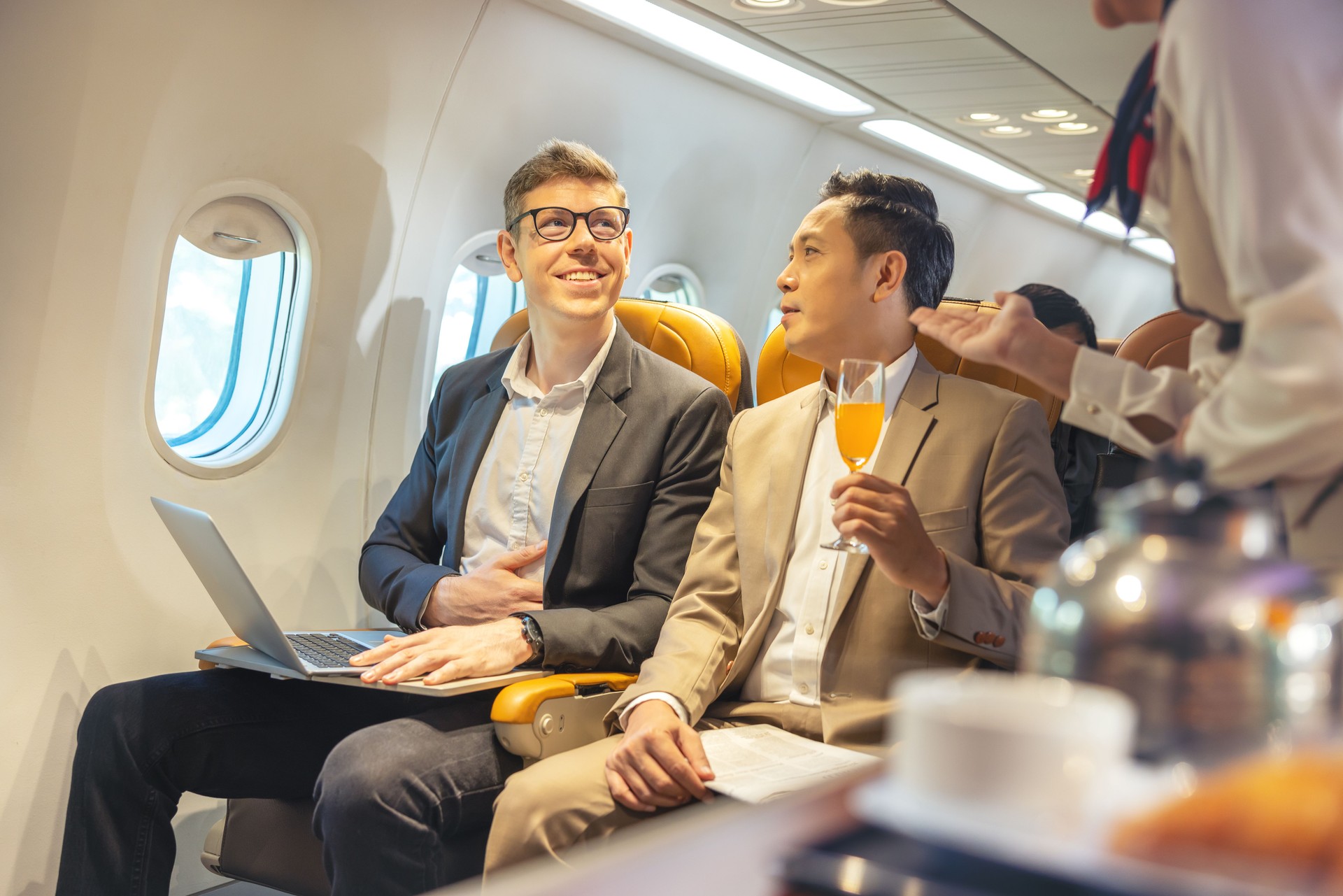 On a corporate aircraft, an air hostess serves passengers. On an airplane, a flight attendant serves a drink to a businessman who is working on his laptop.